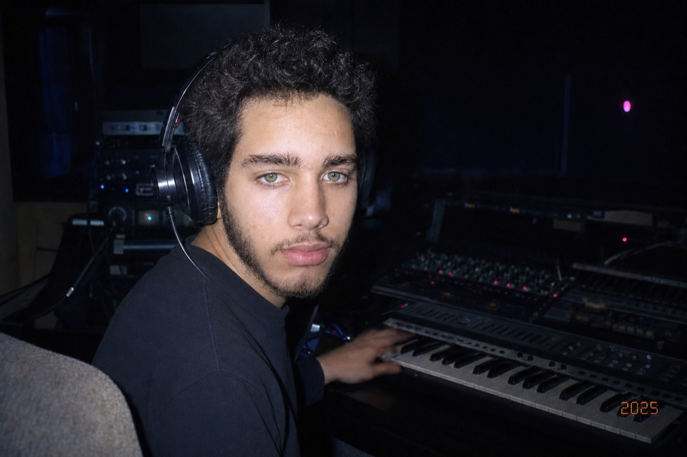 Igor de fones no home studio, mão sobre o teclado MIDI, mesa de mixagem ao fundo, foto com flash datada de 2025.