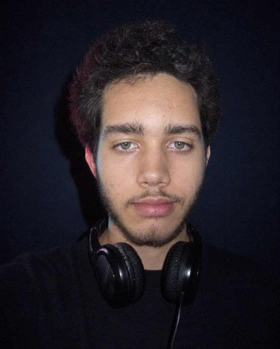 Retrato de Igor Lagioia Freire, o Joya: 18 anos, cabelo cacheado, fones de ouvido no pescoço, fundo escuro com luz rosa e azul.